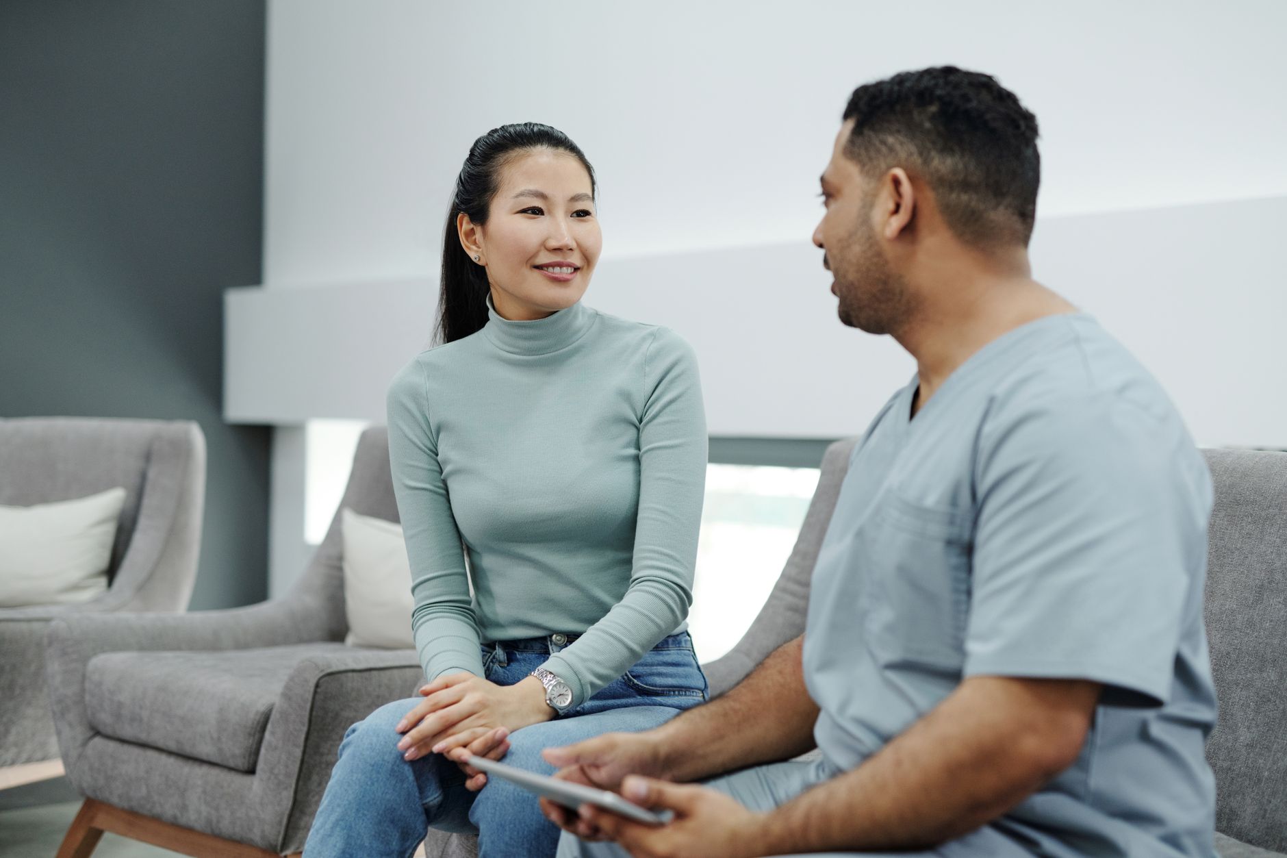 A woman in a sage turtleneck sits on a gray sofa listening to a health care worker in blue scrubs who holds a tablet