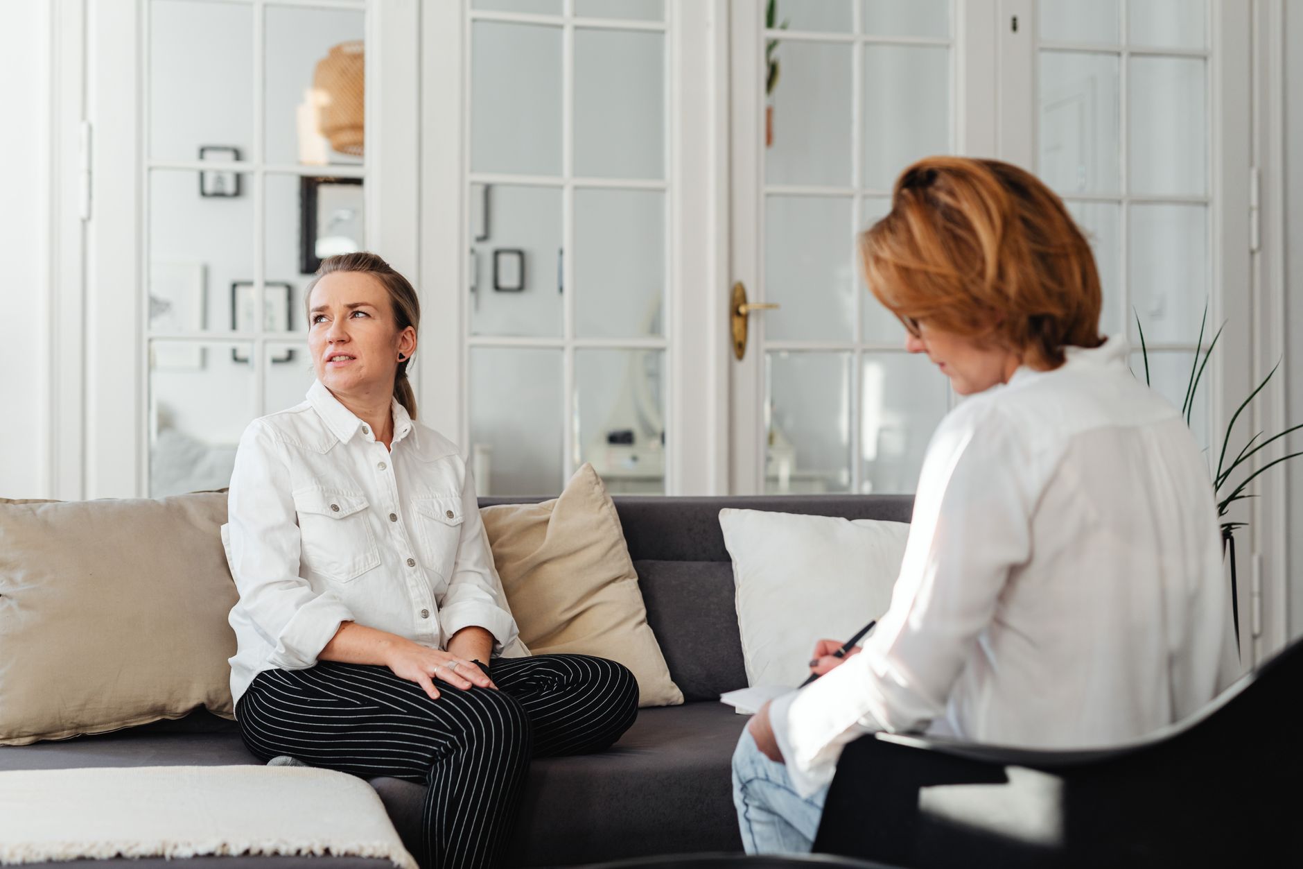 A woman sits on a gray sofa talking while a counselor across from her takes notes in a bright room with glass doors