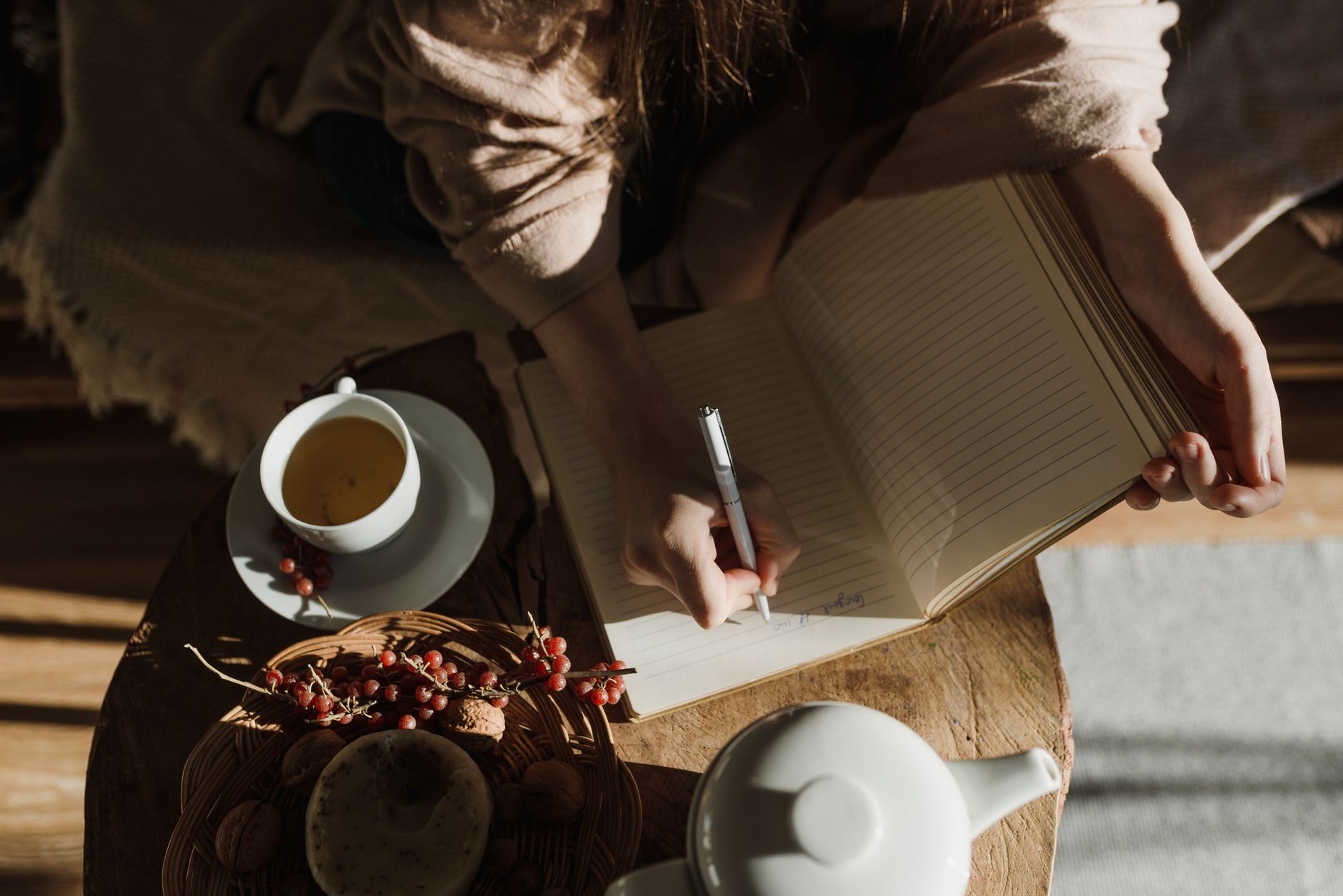 Seen from above, a person in a soft sweater writes in a lined journal beside a cup of tea in warm sunlight