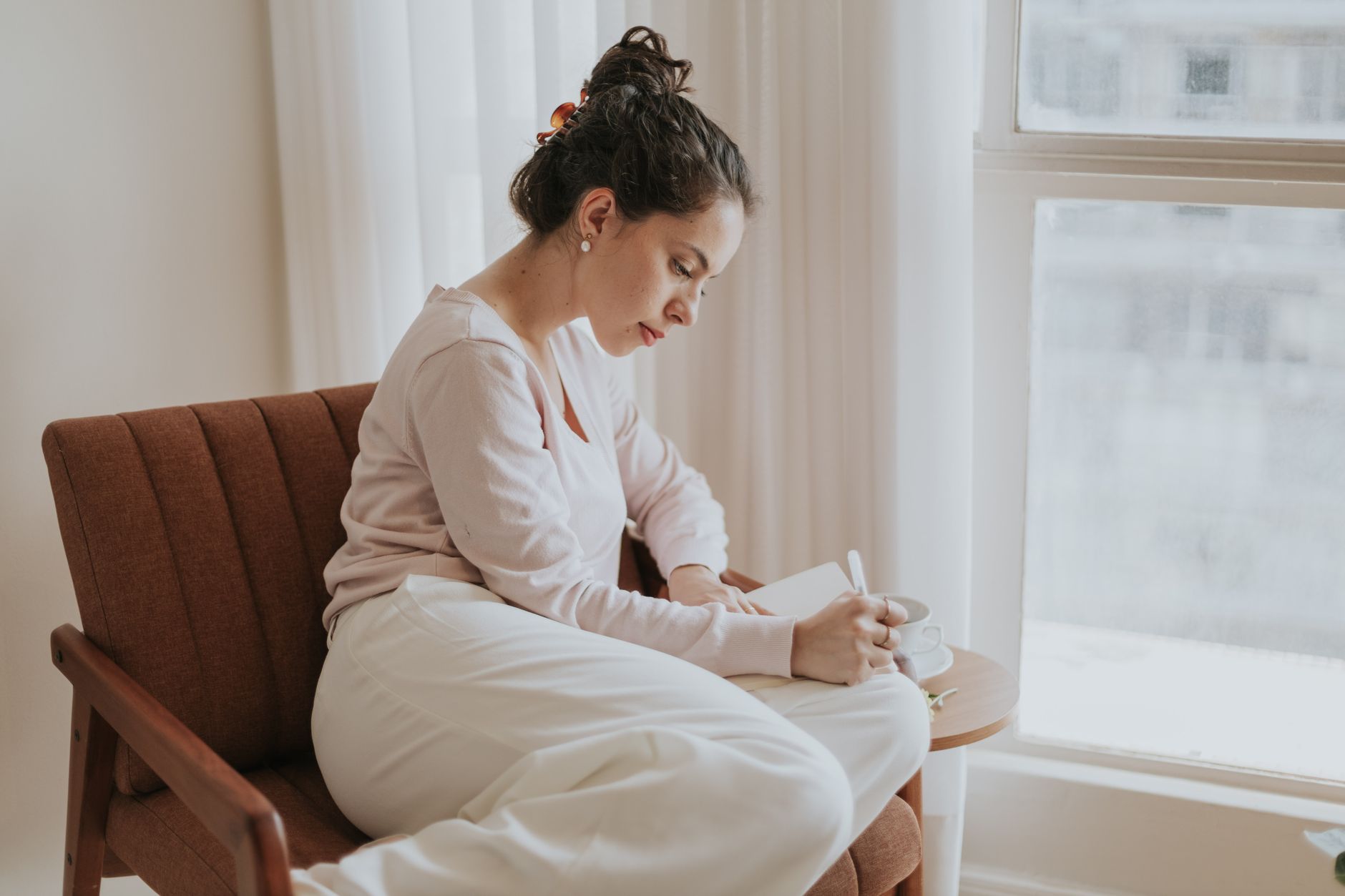 A woman in a pale pink sweater writes in a notebook on a wooden armchair by a bright window, a cup of tea beside her