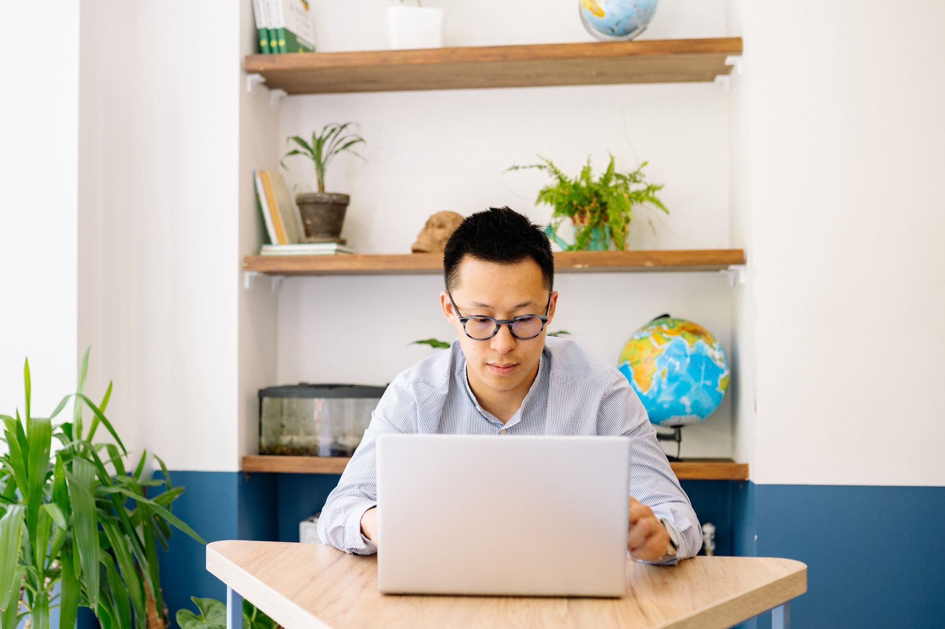 A man in glasses works on a laptop at a wooden desk in a bright room with plants and a globe on the shelves