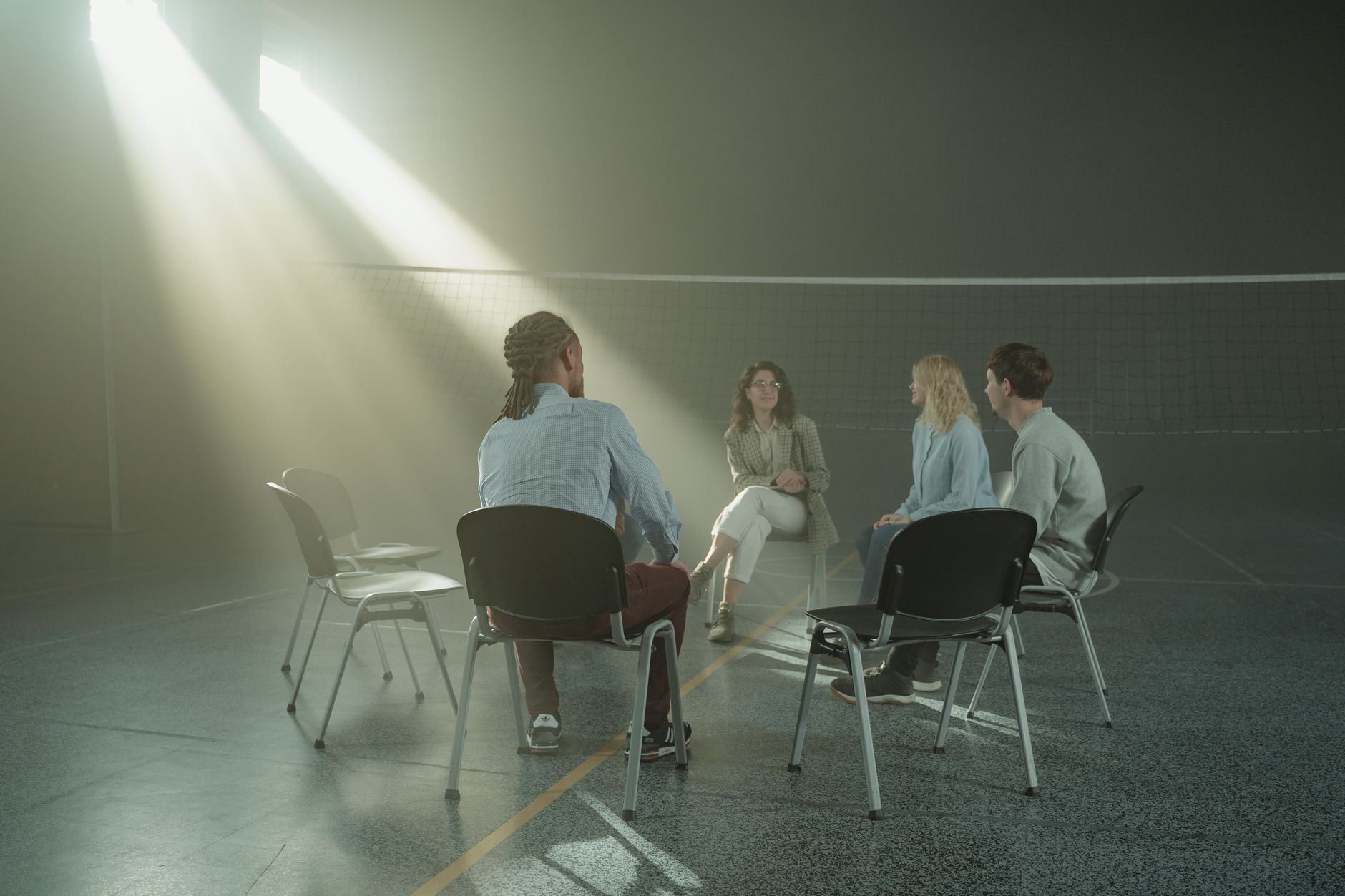 Four adults sit on chairs in a circle talking in a large sunlit hall, with a counselor in a plaid jacket smiling