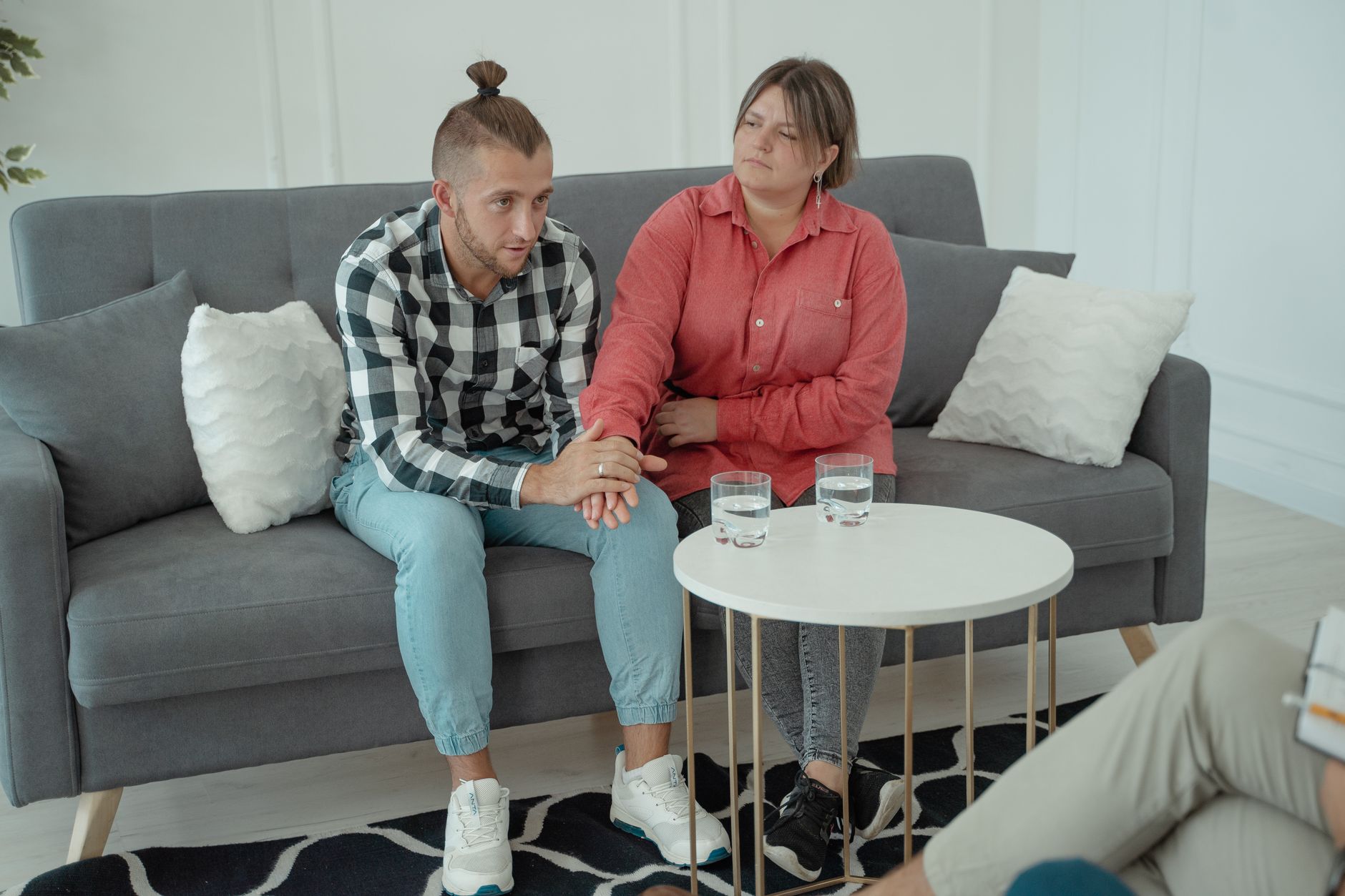 A man and a woman hold hands on a gray sofa while talking with a counselor across a small white table with two glasses of water