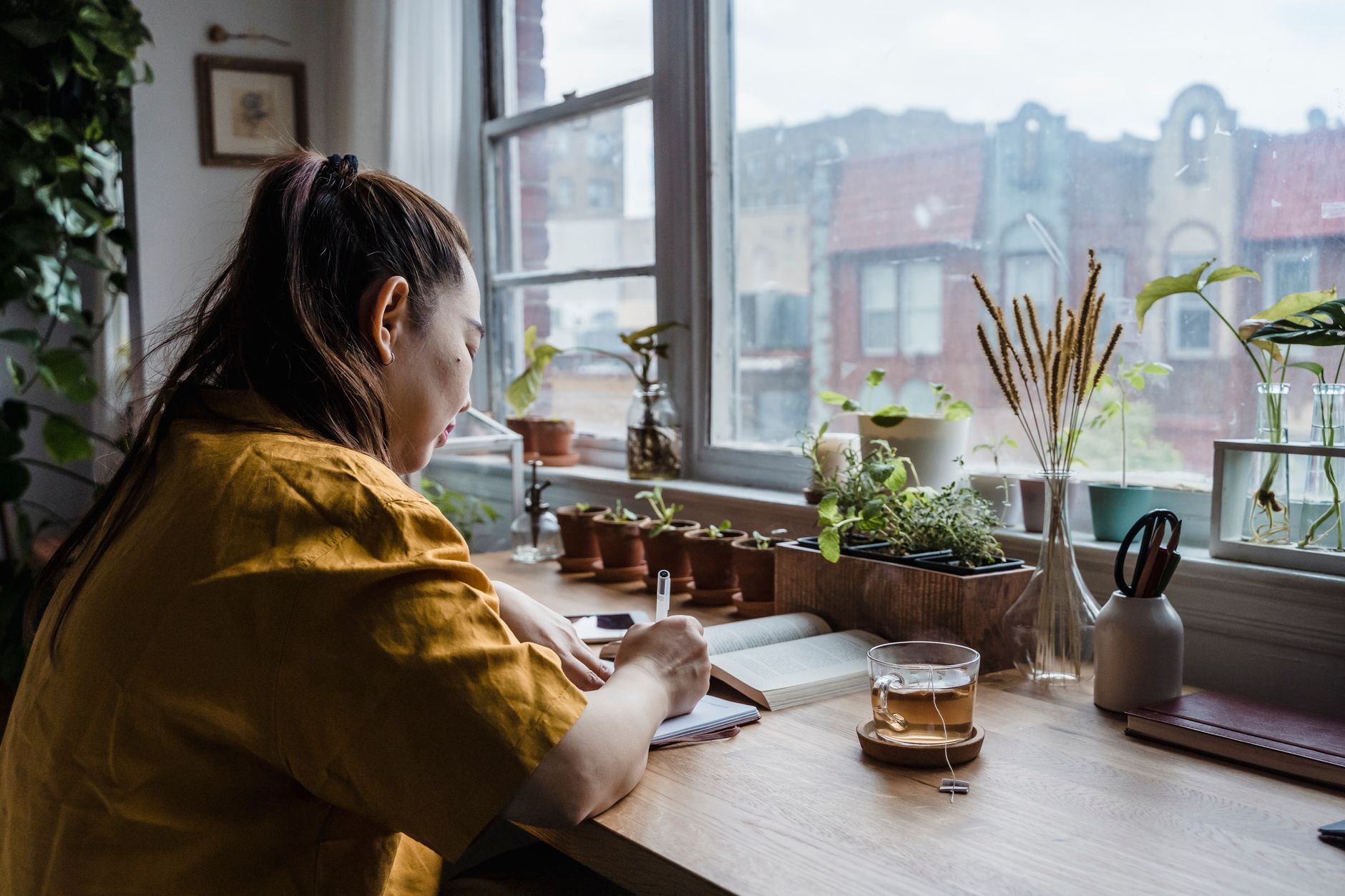 A woman in a mustard shirt writes in a notebook at a wooden desk by a window lined with potted herbs and a glass of tea