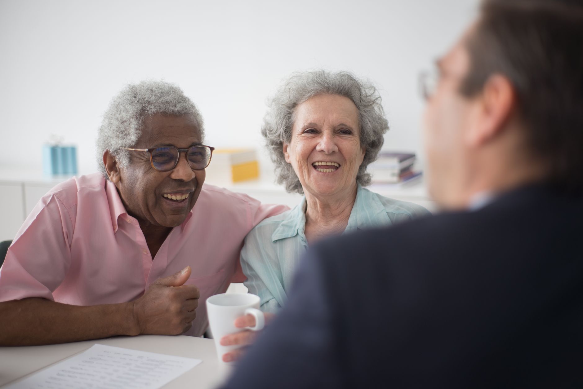 An older couple with gray hair smile and talk with a professional across a table, the woman holding a white mug