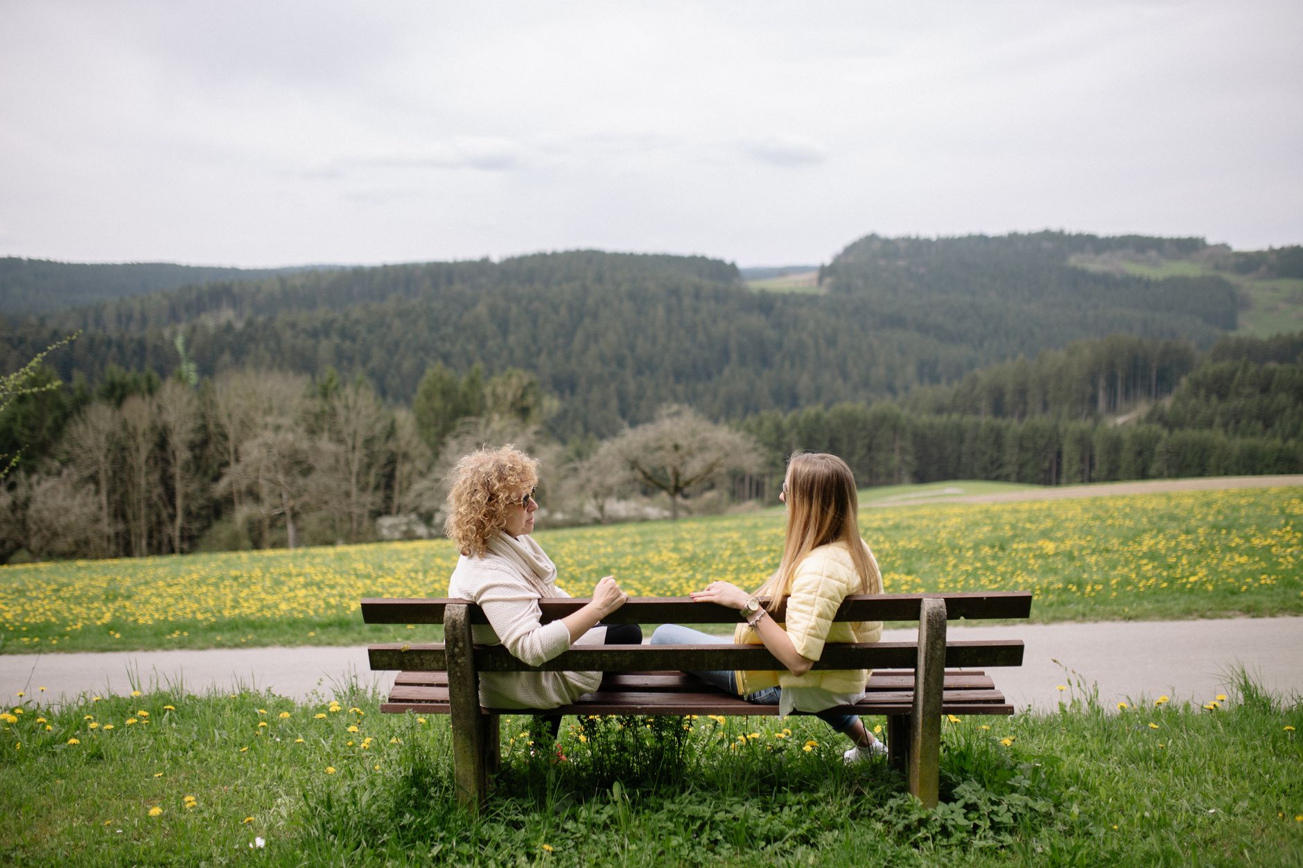 Two women sit on a wooden bench facing each other in conversation, with a dandelion meadow and forested hills behind them