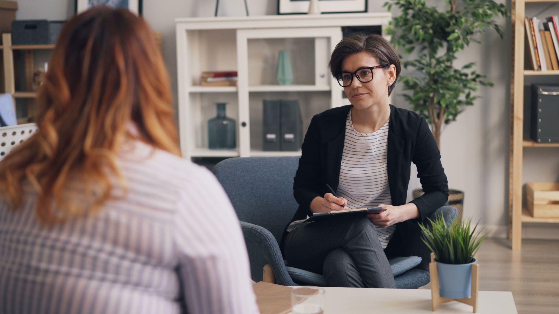 A therapist with glasses and a clipboard listens to a client in a bright office with shelves and potted plants