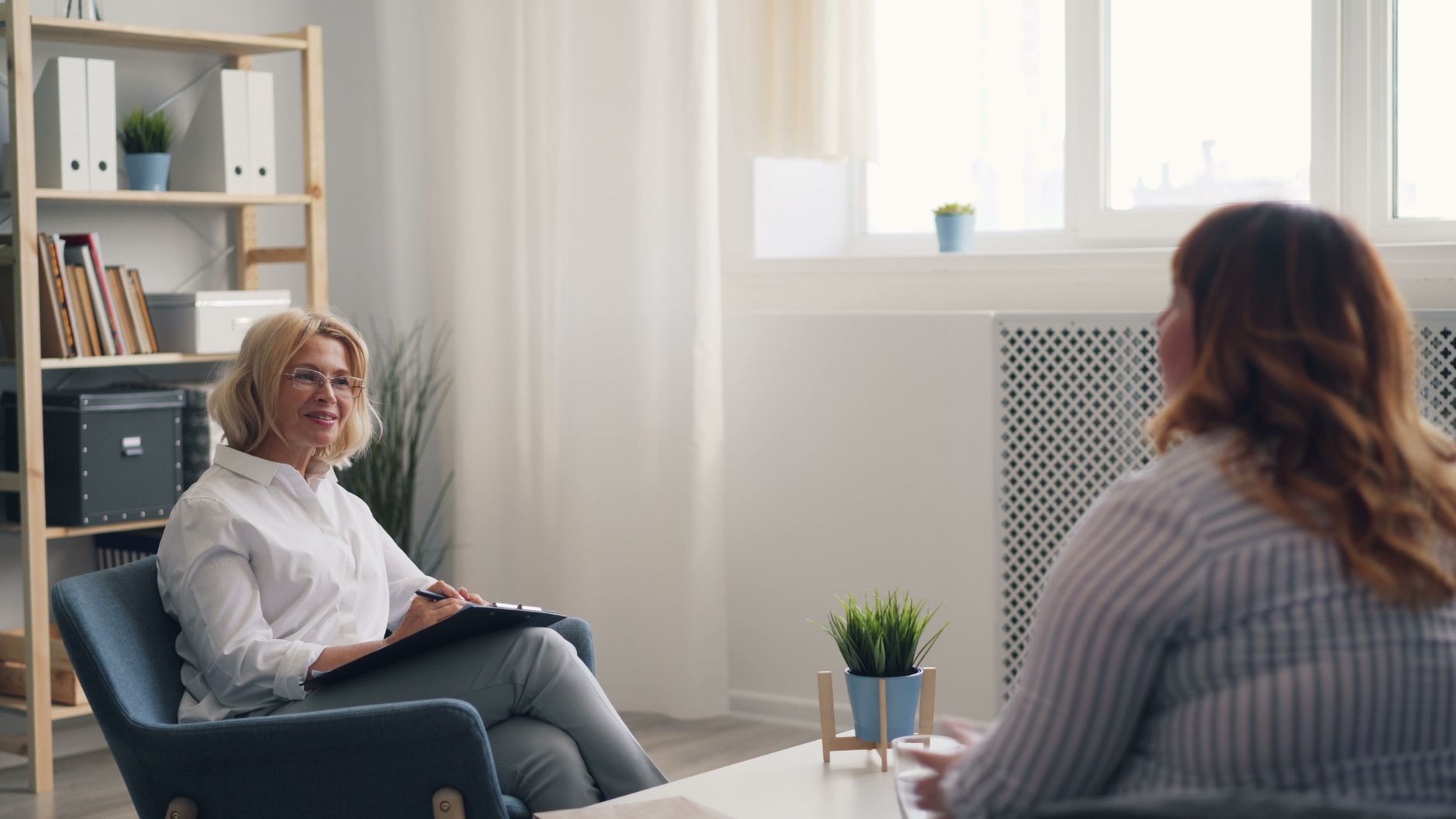 A counselor with a clipboard smiles as she listens to a client seated across from her in a bright room with plants and a bookshelf