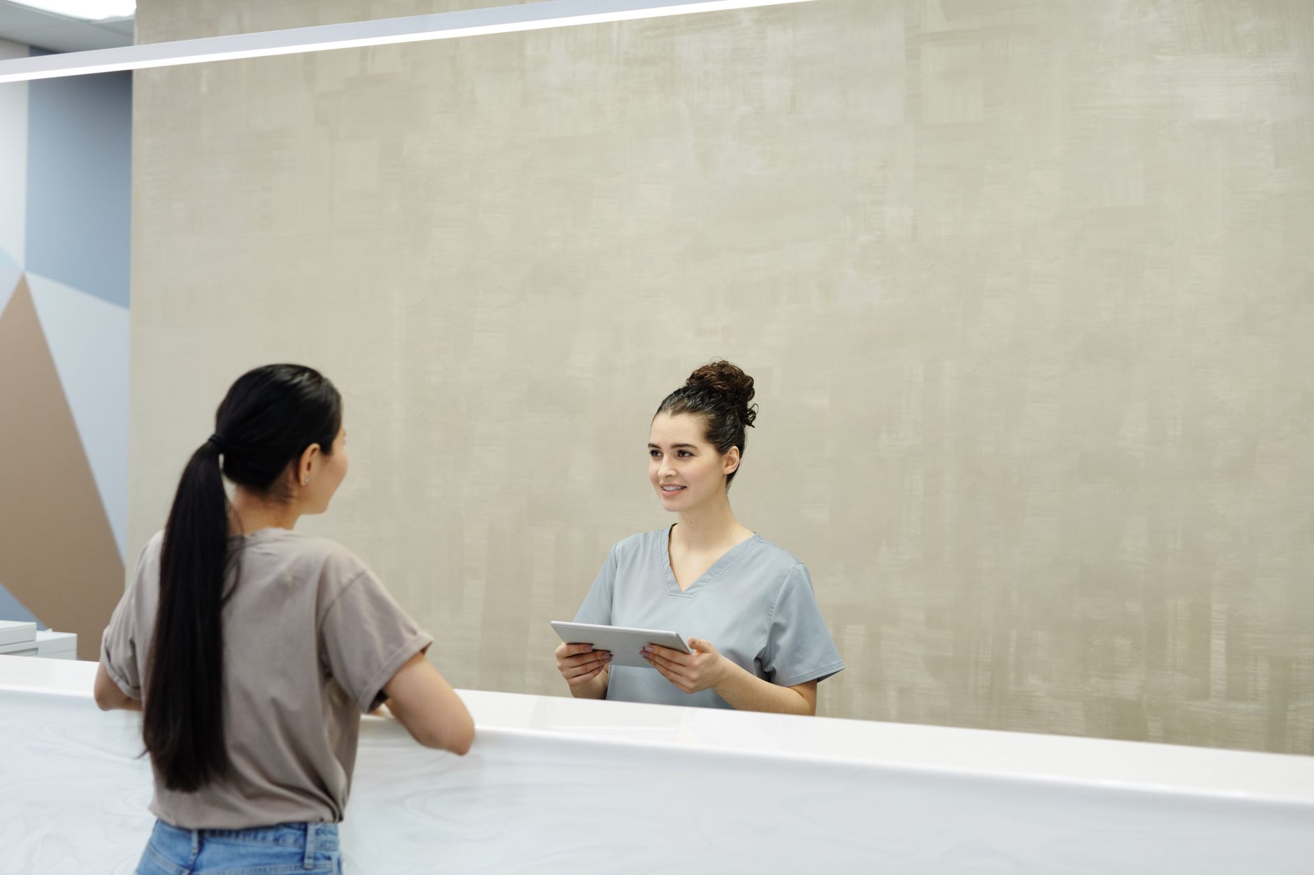 A woman with a long ponytail talks with a smiling clinic receptionist who holds a tablet at a white front desk