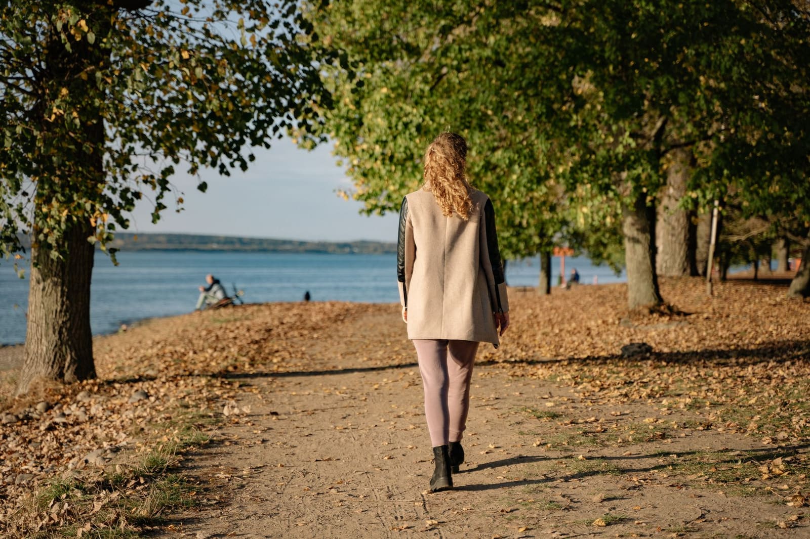 A woman in a beige coat walks alone along a sunny lakeside path covered in fallen autumn leaves