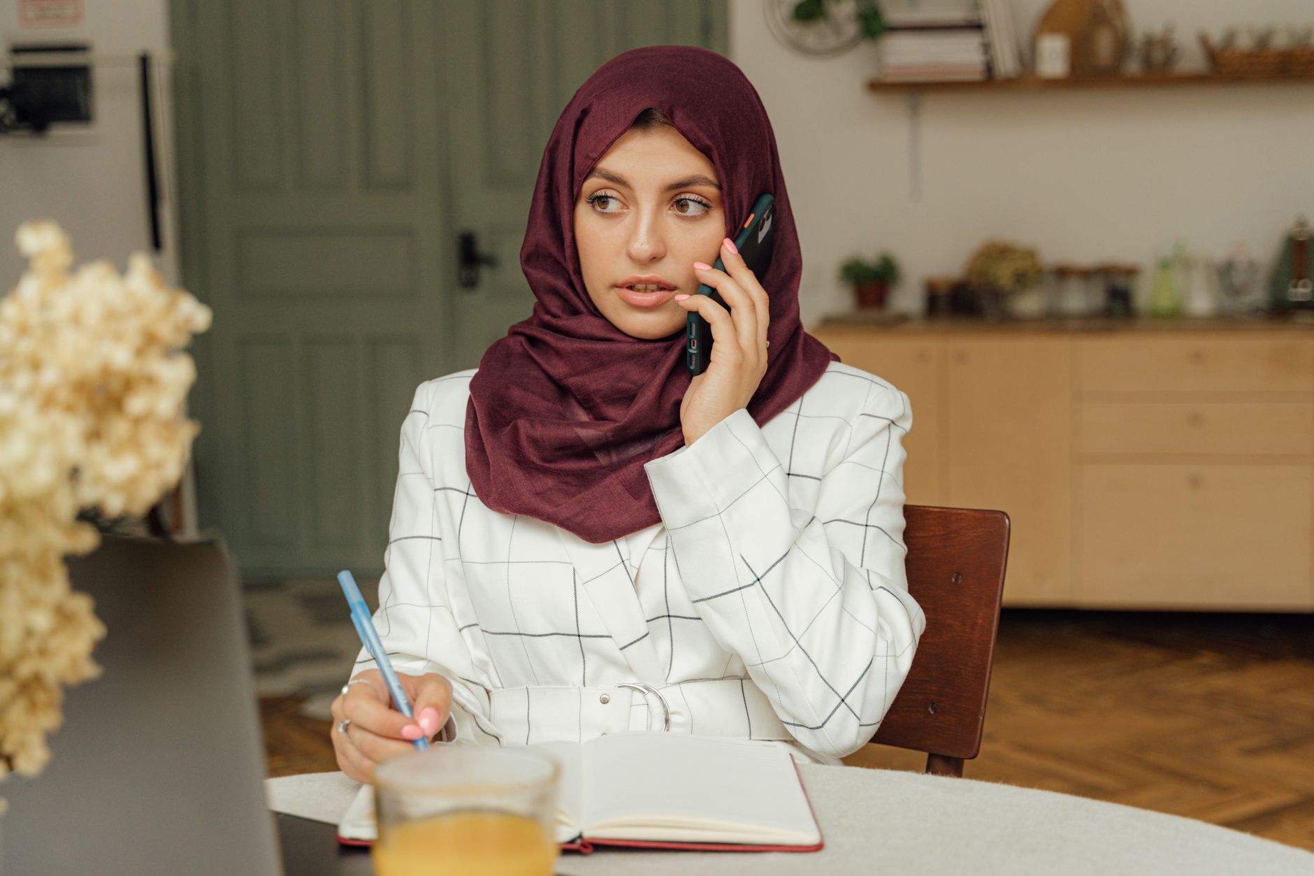 A woman in a headscarf talks on her phone at a kitchen table while writing notes in an open notebook