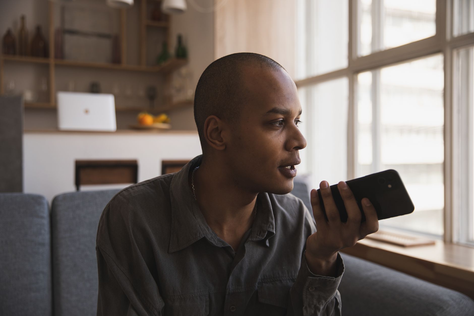 A man sits on a gray sofa by a large window, speaking into a phone held in front of him