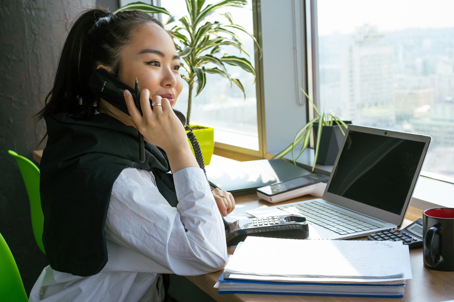 A young woman smiles while talking on a desk phone beside a laptop, a stack of folders and a mug by a sunny office window