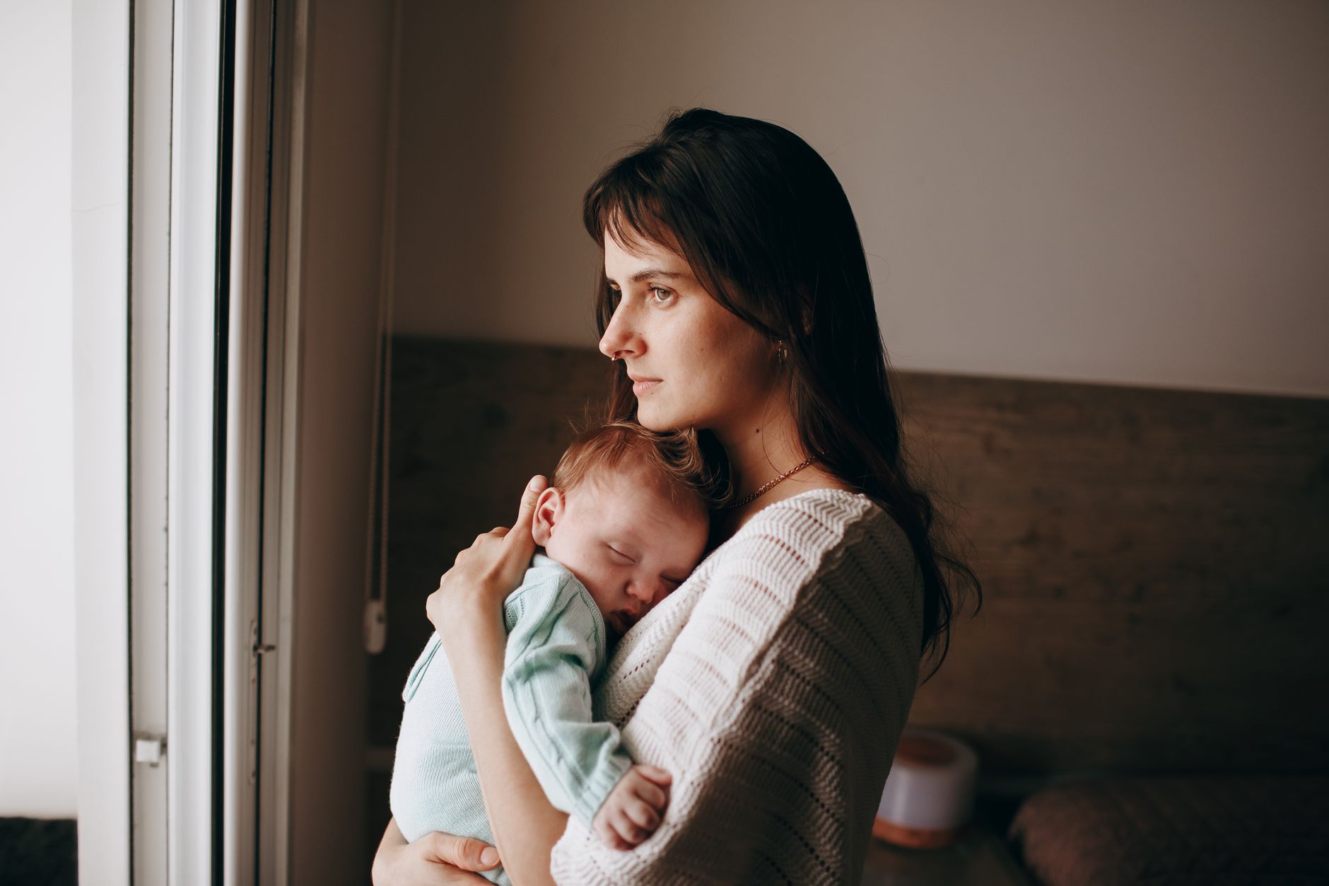 A young mother in a cream knit sweater stands by a window holding her sleeping newborn against her shoulder