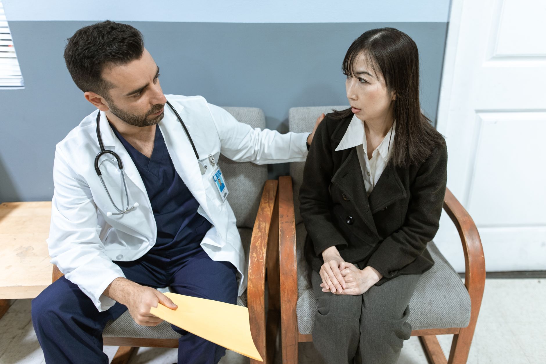 A doctor in a white coat sits beside a woman in a clinic waiting area and talks with her while holding a folder