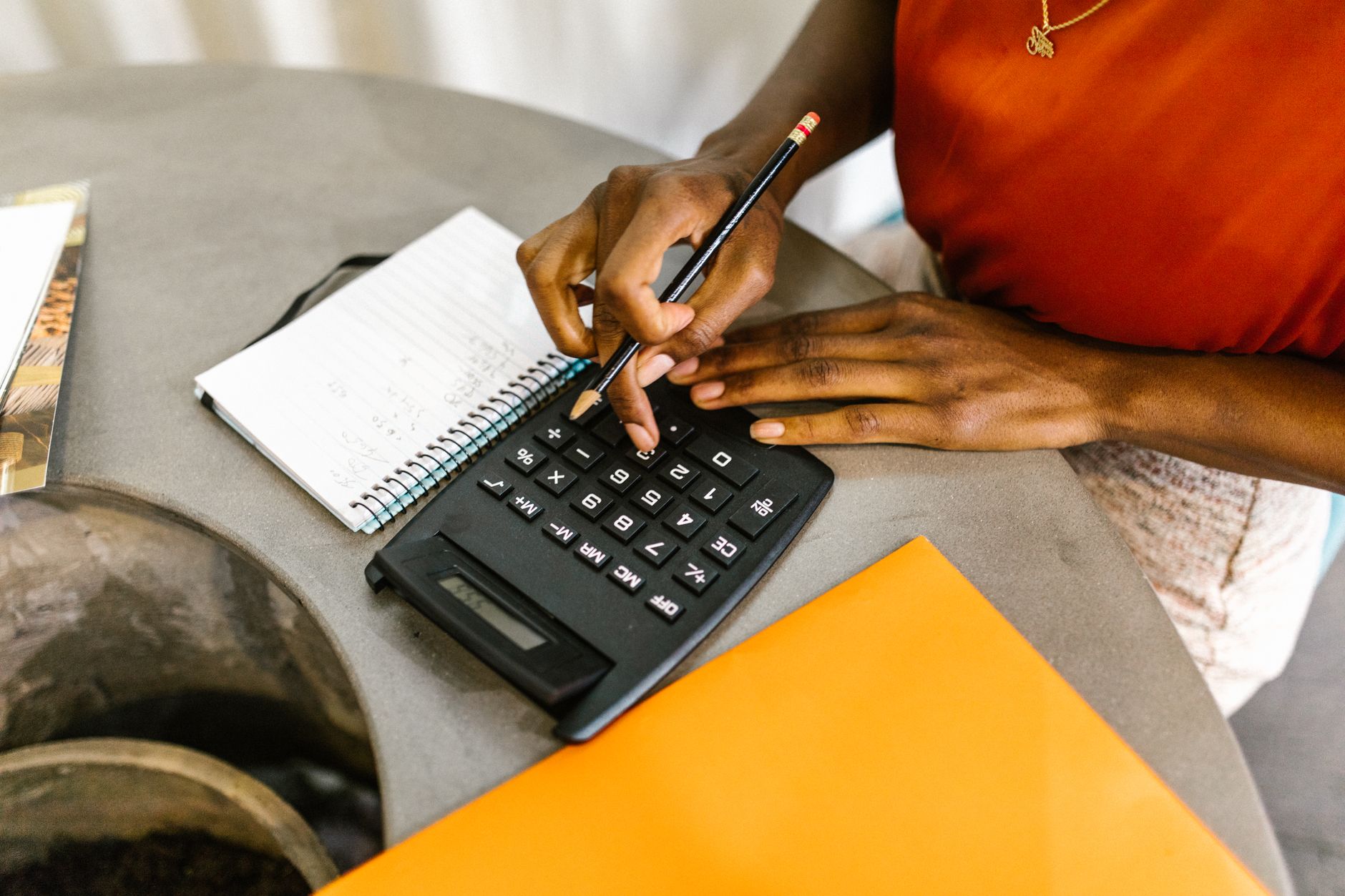 A woman in an orange top works out figures on a black calculator with a pencil next to a spiral notebook