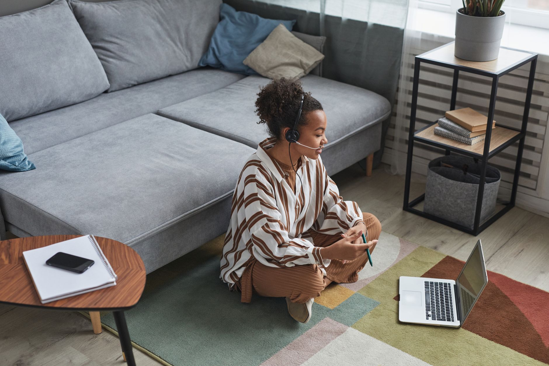 A woman wearing a headset sits cross-legged on a living room rug, talking to someone on a laptop in front of a gray sofa