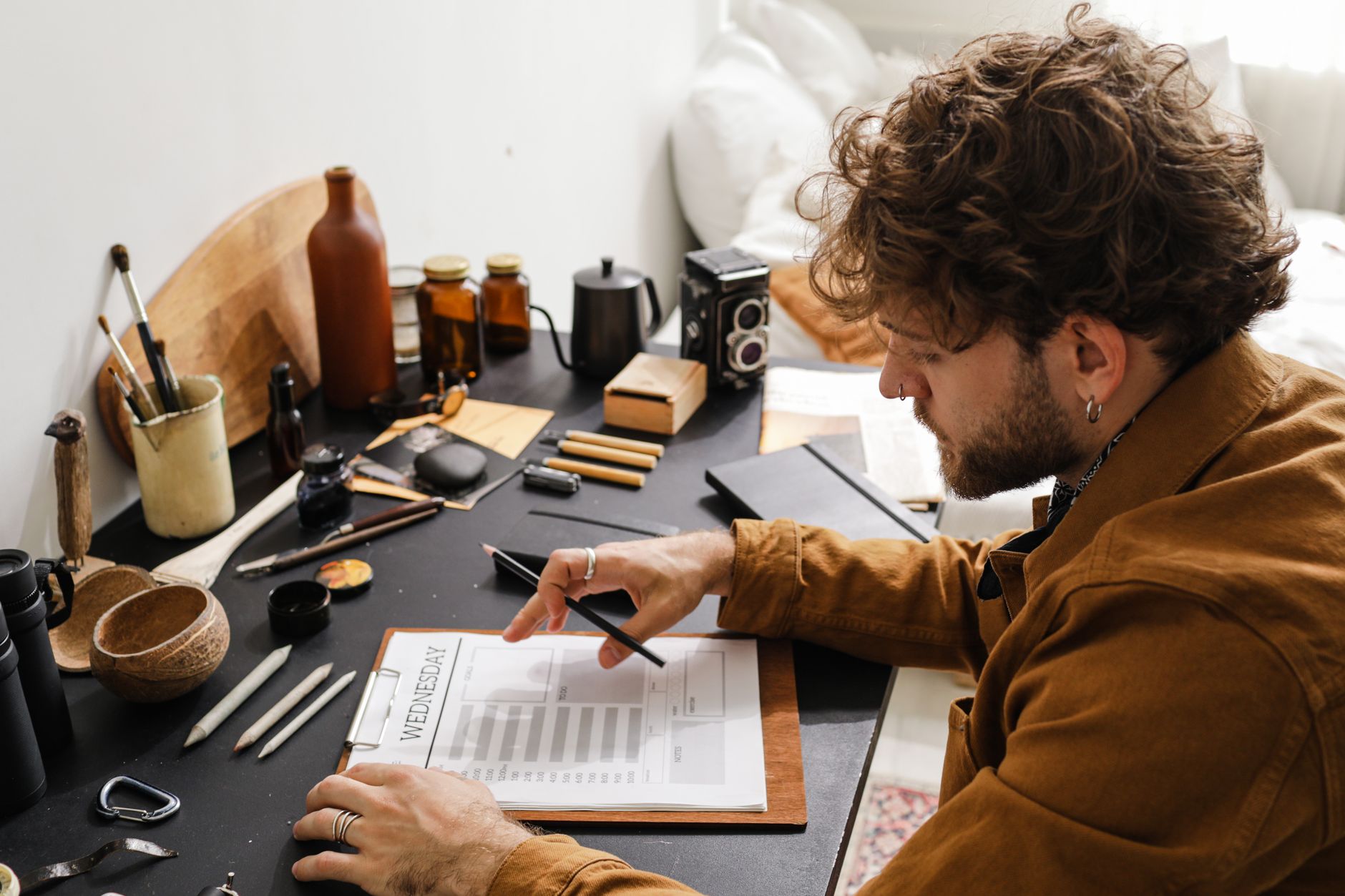 A man with curly hair in a mustard jacket fills in a daily planner on a clipboard at a cluttered desk