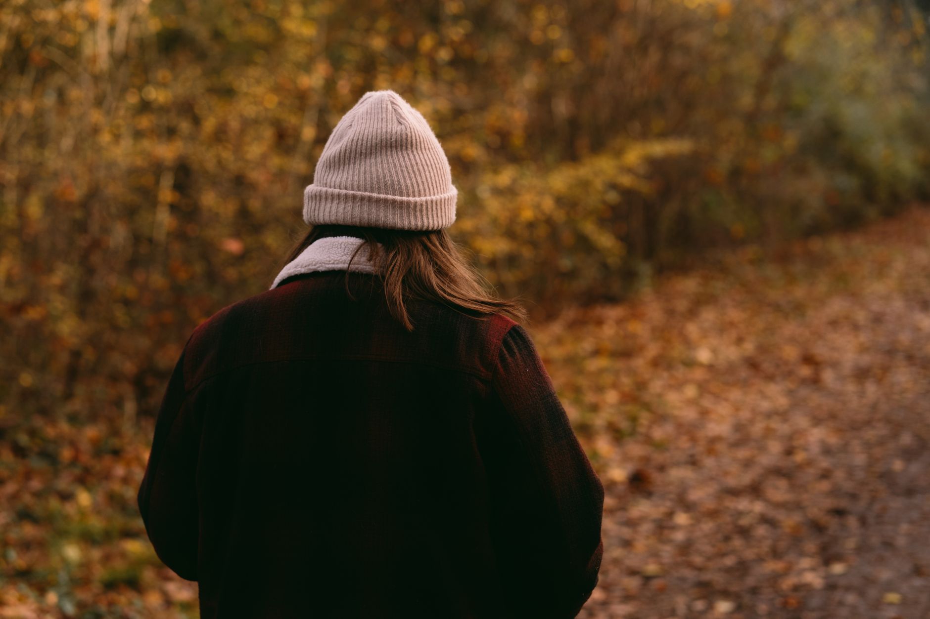 A person in a knit beanie and dark wool coat walks along a leaf-covered path through autumn woods, seen from behind