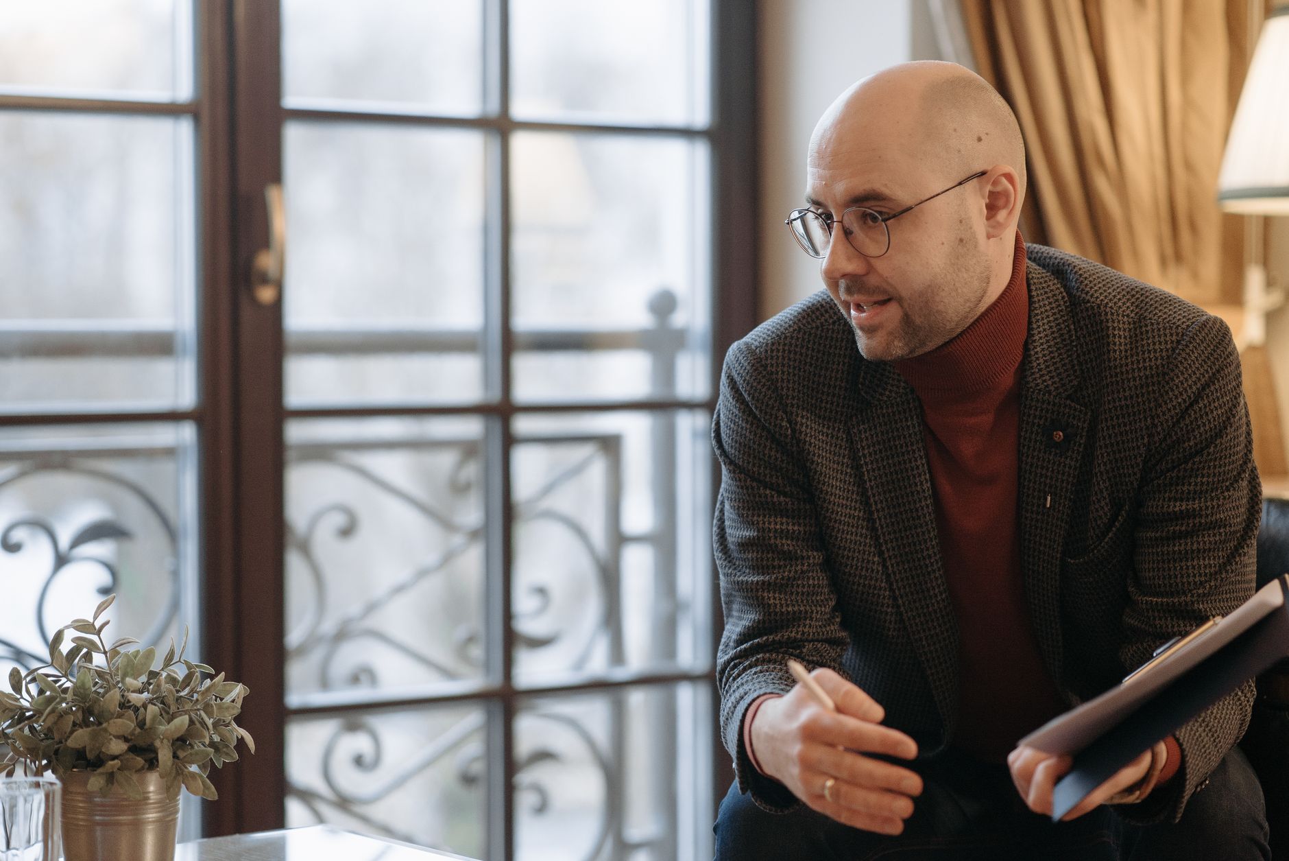 A therapist in glasses and a tweed jacket leans forward, pen in hand, listening during a session by a tall window