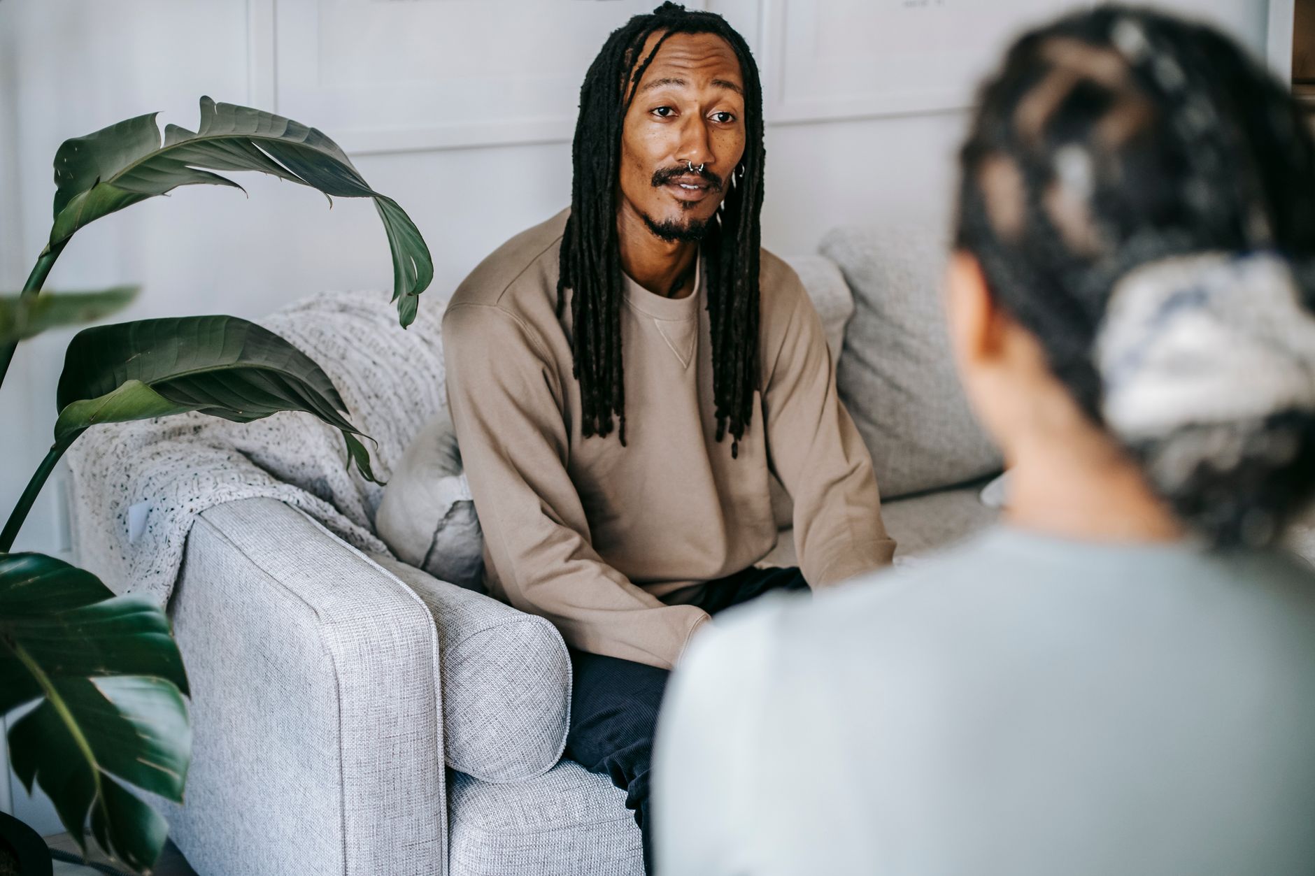 A man in a beige sweatshirt sits on a light gray sofa beside a large plant, talking with a counselor seen from behind