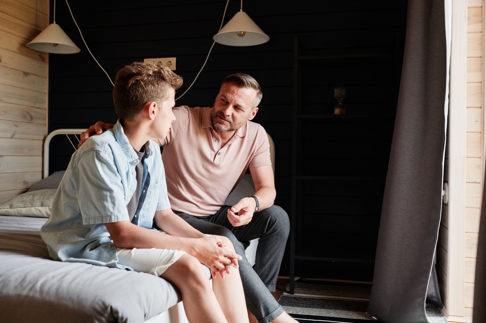 A father in a pink polo shirt sits on a bed with his arm around his teenage son, talking quietly in a wood-paneled bedroom
