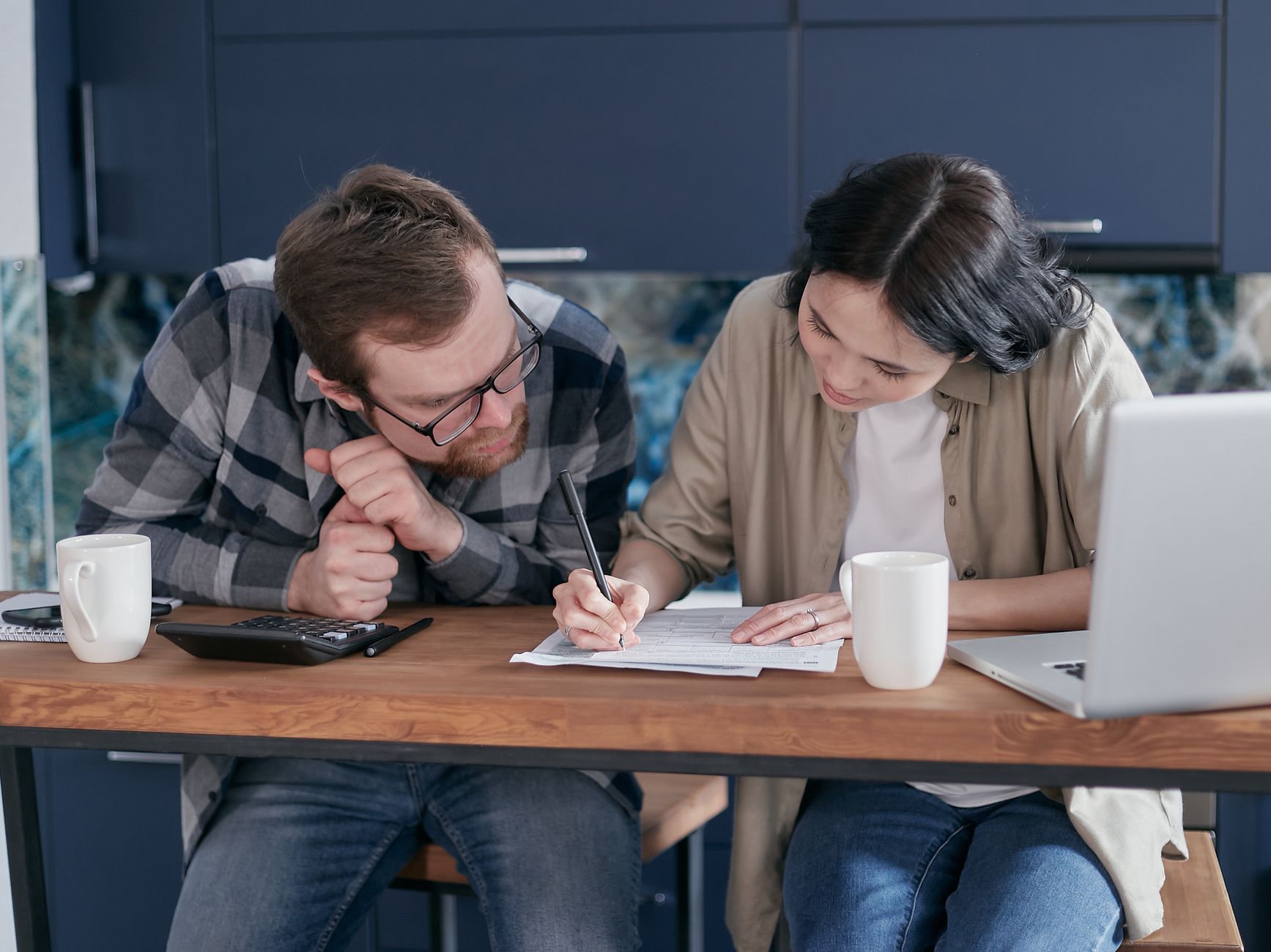 A man in glasses and a plaid shirt watches a woman write on a form at a wooden kitchen table with a calculator, mugs and a laptop